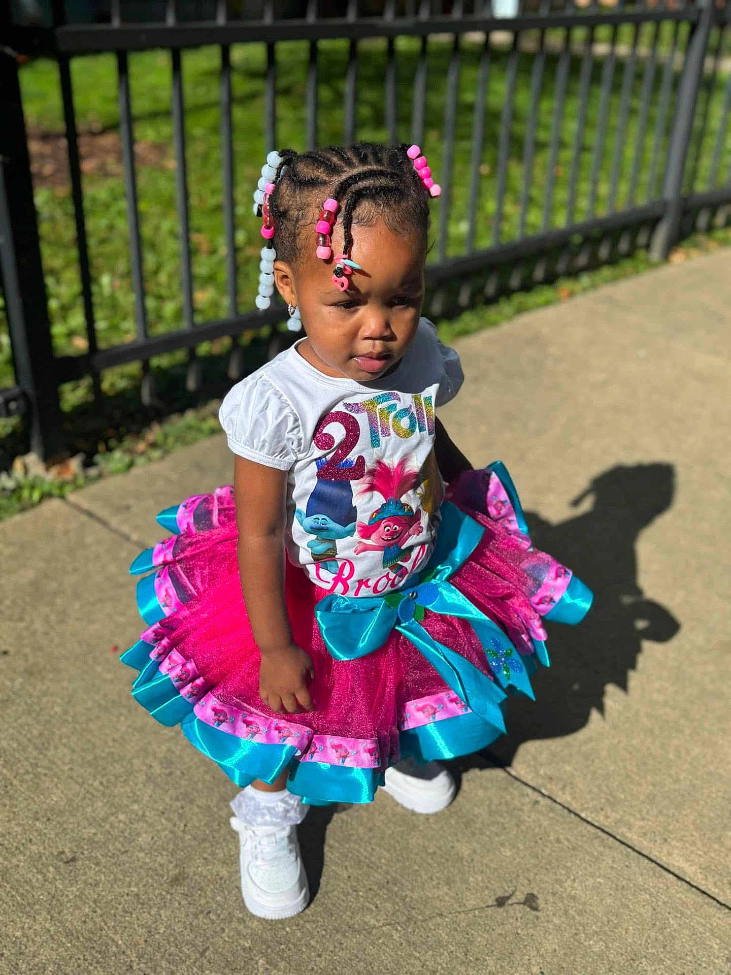 Brooklyn joined the competition — help win amazing prizes! child, toddler, girl, outdoor, sunlight, tutu, braids, beads, trolls, shirt, white_sneakers, sidewalk, shadow, fence, greenery, colorful, fashion, cute, portrait, standing