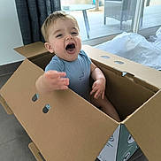 Elio participe au concours pour gagner de l'argent avec cette photo : toddler, child, cardboard_box, indoor, floor, window, glass_door, curtain, smiling, happy, short_hair, blue_clothing, playful, mouth_open, furniture, table, chair, natural_light, home, person