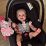 baby, infant, car_seat, maxi_cosi, toy, pink, floral, floor, wooden_floor, happy, smiling, face, feet, straps, sitting, indoor, child, person, cute, playful