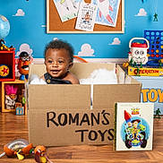 Roman joined the competition — help win amazing prizes! toddler, child, baby, portrait, smiling, sitting, cardboard_box, toy_box, toys, playroom, colorful, indoors, face, curly_hair, overalls, wooden_floor, books, robot_toy, board_game, stuffed_toy
