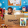 toddler, child, baby, portrait, smiling, sitting, cardboard_box, toy_box, toys, playroom, colorful, indoors, face, curly_hair, overalls, wooden_floor, books, robot_toy, board_game, stuffed_toy