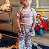 child, toddler, standing, pacifier, portrait, indoors, living_room, couch, toy, toy_walker, colorful, floral_clothing, pants, shirt, rug, furniture, window, air_conditioner,  curious, family_home