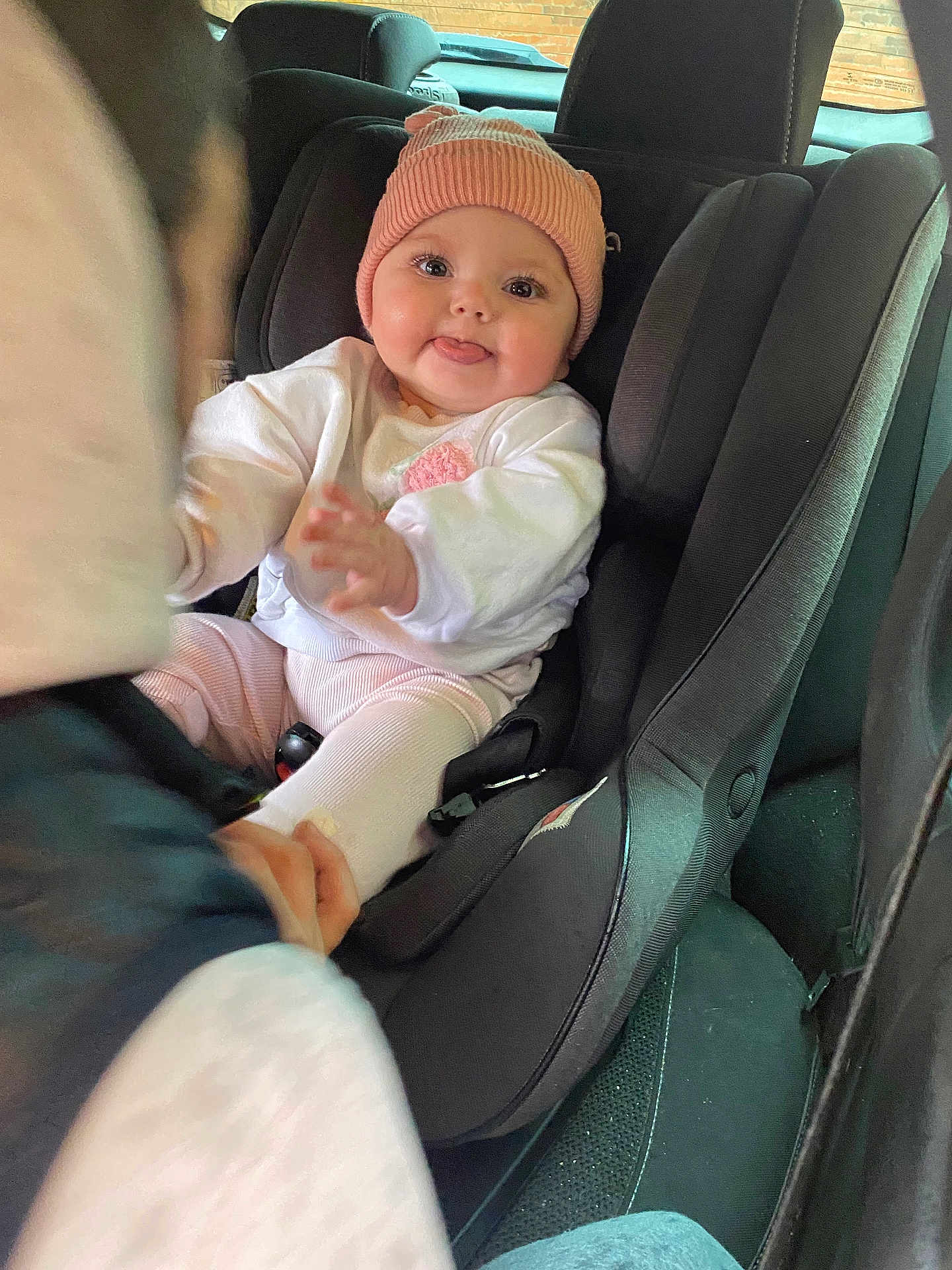 Bonnie is registered to the contest to win money with this photo: baby, car_seat, child, pink_hat, white_clothing, tongue_out, smiling, car_interior, seatbelt, hand, cute, infant, person, portrait, sitting, warm_clothing, playful, young_child, car, seat