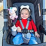 toddler, child, stroller, headband, red_shirt, blue_jeans, unicorn_toy, bottle, happy, smiling, curly_hair, outdoor, seatbelt, baby, person, cute, daylight, casual_clothing, playful, comfort