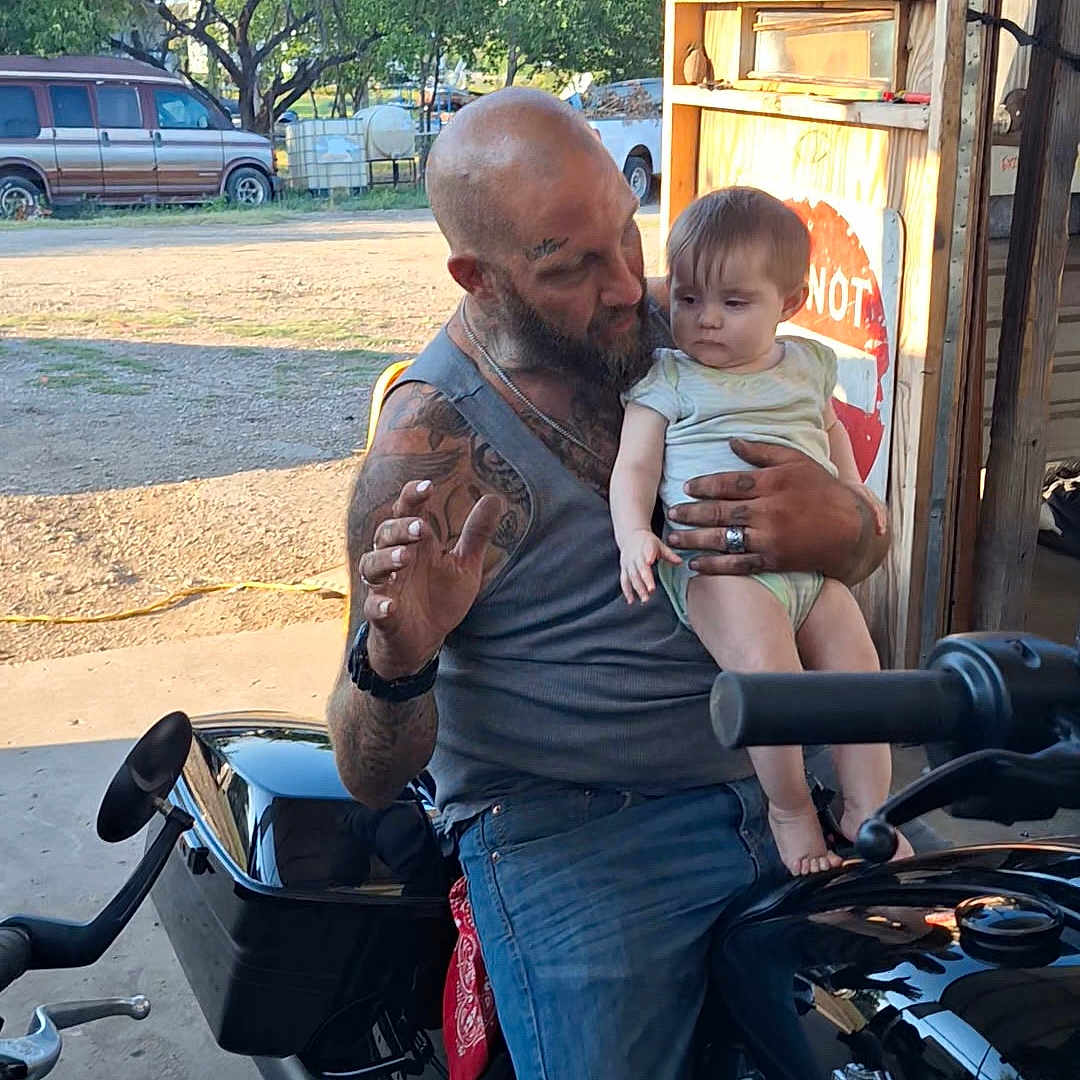 Harley is registered to the contest to win money with this photo: baby, beard, casual, child, dirt, garage, handlebars, jeans, man, motorcycle, outdoor, person, sign, sunlight, tank_top, tattoos, trees, van, vehicle, wood