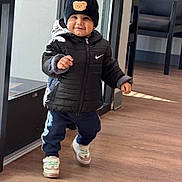 Aryan is registered to the contest to win money with this photo: toddler, child, indoor, walking, black_jacket, beanie, hat, sneakers, floor, window, door, smiling, cute, clothing, person, young_child, happy, pants, footwear, headwear