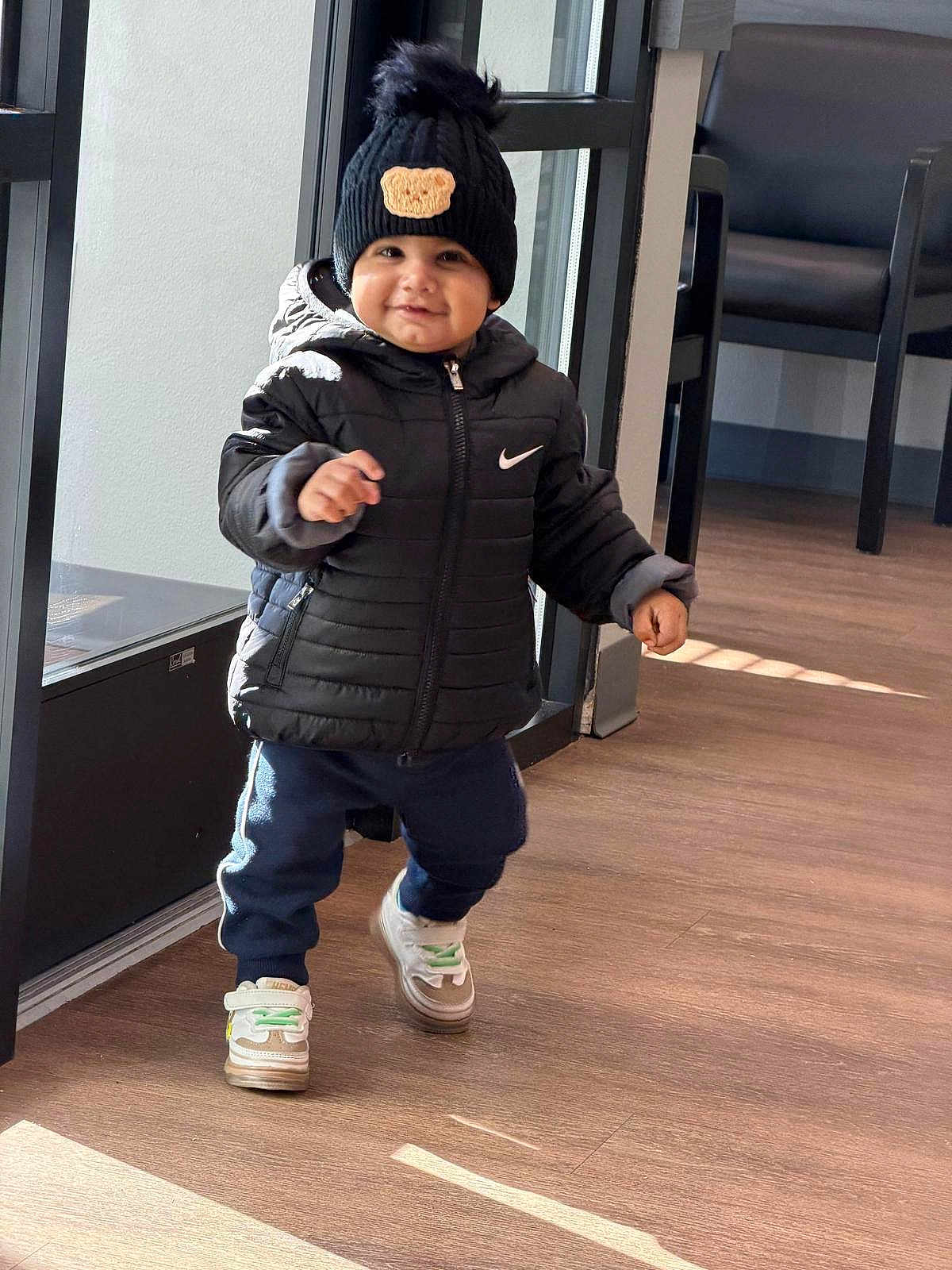 Aryan is registered to the contest to win money with this photo: toddler, child, indoor, walking, black_jacket, beanie, hat, sneakers, floor, window, door, smiling, cute, clothing, person, young_child, happy, pants, footwear, headwear
