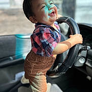 Zaid is registered to the contest to win money with this photo: toddler, child, smiling, steering_wheel, car_interior, plaid_shirt, corduroy_pants, happy, standing, baby, vehicle, seat, hand, portrait, cute, young_child, indoor, clothing, sunlight, playful