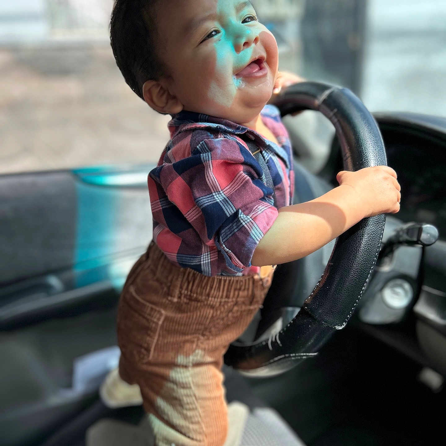 Zaid is registered to the contest to win money with this photo: baby, car_interior, child, clothing, corduroy_pants, cute, hand, happy, indoor, plaid_shirt, playful, portrait, seat, smiling, standing, steering_wheel, sunlight, toddler, vehicle, young_child