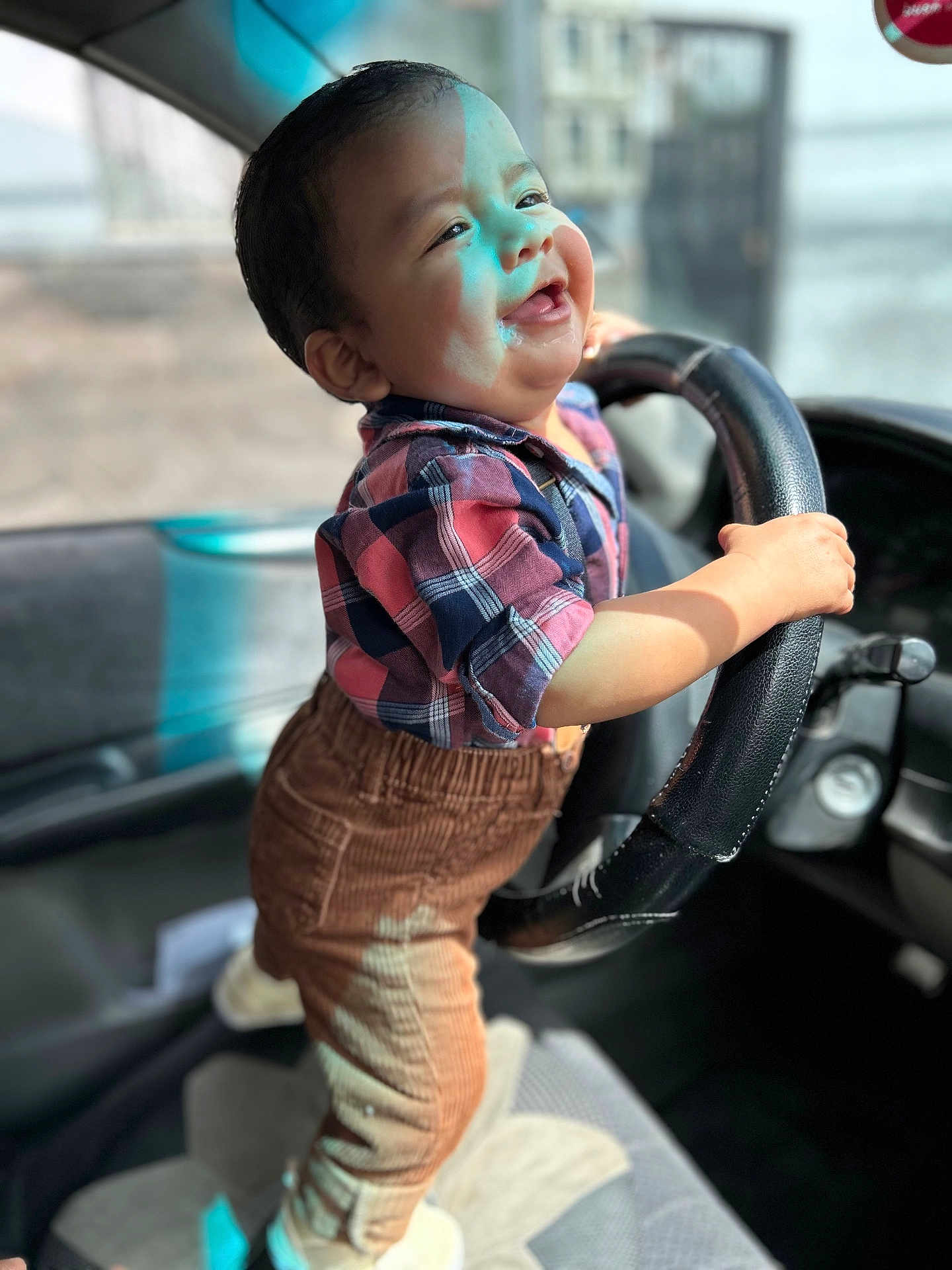 Zaid is registered to the contest to win money with this photo: toddler, child, smiling, steering_wheel, car_interior, plaid_shirt, corduroy_pants, happy, standing, baby, vehicle, seat, hand, portrait, cute, young_child, indoor, clothing, sunlight, playful