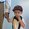 child, boy, cap, suspenders, white_shirt, brown_pants, adult_hand, holding_hand, indoor, wall, decor, spider_web, text_overlay, casual, portrait, standing, young, curious, fashion, people