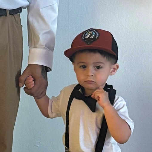 Julian is registered to the contest to win money with this photo: child, boy, cap, suspenders, white_shirt, brown_pants, adult_hand, holding_hand, indoor, wall, decor, spider_web, text_overlay, casual, portrait, standing, young, curious, fashion, people