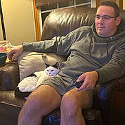 Tiki is registered to the contest to win money with this photo: man, cat, armchair, remote_control, lamp, window, curtains, couch, gray_hoodie, shorts, indoor, relaxed, pet, leather, black_and_white_cat, home, seated, cozy, living_room, casual