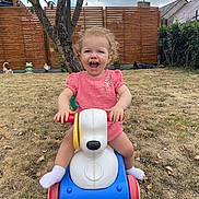 Mya participe au concours pour gagner de l'argent avec cette photo : toddler, child, toy, ride_on_toy, outdoor, grass, garden, tree, fence, smiling, happy, pink_dress, curly_hair, playtime, plastic, yard, daylight, fun, cute, person