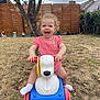 toddler, child, toy, ride_on_toy, outdoor, grass, garden, tree, fence, smiling, happy, pink_dress, curly_hair, playtime, plastic, yard, daylight, fun, cute, person