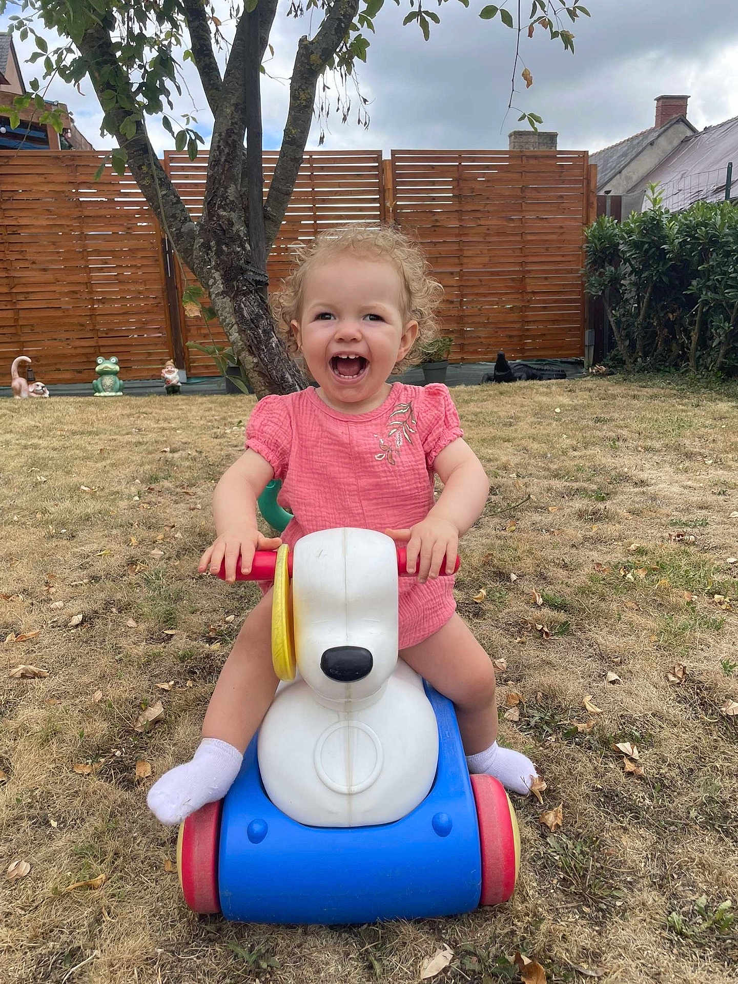 Mya participe au concours pour gagner de l'argent avec cette photo : toddler, child, toy, ride_on_toy, outdoor, grass, garden, tree, fence, smiling, happy, pink_dress, curly_hair, playtime, plastic, yard, daylight, fun, cute, person