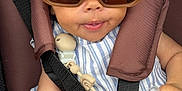 Eliana is registered to the contest to win money with this photo: baby, sunglasses, headband, car_seat, pacifier, clip, striped_clothing, hand, face, child, infant, safety_harness, brown, white, cute, smile, tongue, accessory, seatbelt, closeup