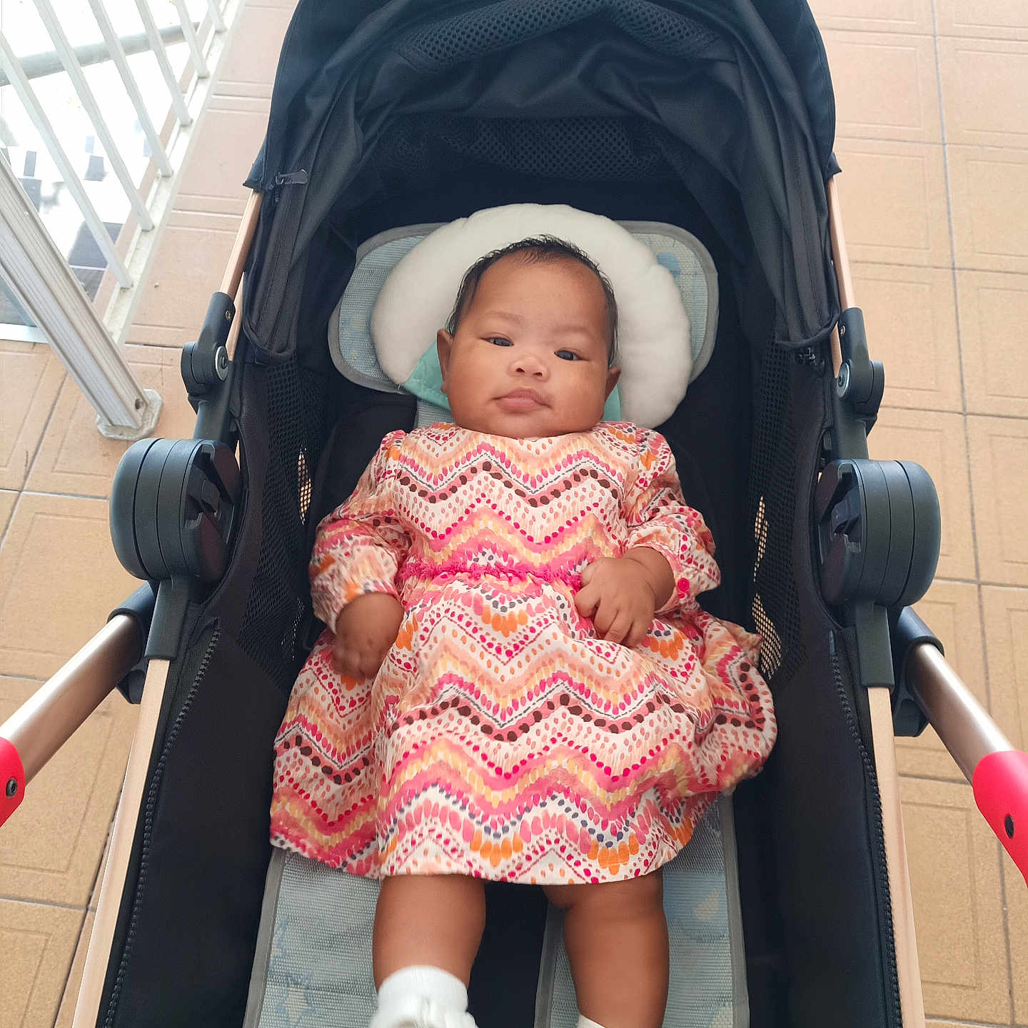 Goreth is registered to the contest to win money with this photo: baby, black, child, cushion, cute, dress, expression, floor_tiles, footwear, headrest, indoor, infant, patterned_clothing, person, portrait, railing, seated, socks, stroller, young