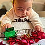 baby, child, face, hands, sweater, gift_bow, red_bow, green_bow, white_bow, holiday, christmas, curious, closeup, indoor, soft_light, table, decorations, focus, portrait, candid