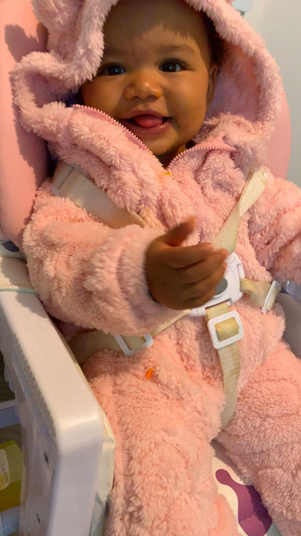 Daïanna participe au concours pour gagner de l'argent avec cette photo : baby, child, pink_onesie, fluffy, smiling, tongue_out, high_chair, safety_straps, cute, indoor, happy, person, infant, seat, clothing, face, hood, hand, closeup, portrait