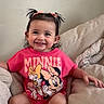 child, toddler, smiling, sitting, pink_shirt, minnie_mouse, pigtails, couch, cozy, blanket, indoors, cute, happy, person, face, hair_bow, white_socks, casual, comfortable, portrait
