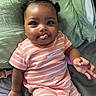 baby, infant, child, striped_clothing, peach_color, tongue_out, curly_hair, hair_buns, bed, bedding, green_sheet, gray_sheet, hand, cute, playful, lying_down, person, portrait, indoors, smile