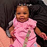 baby, child, smiling, pink_clothing, pacifier_clip, bed, blanket, patterned_sheet, cute, happy, indoors, person, portrait, infant, braided_hair, earrings, decorative_clothing, seated, black_blanket, colorful