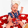american_flag, baby, blue, bow, child, clothing, cute, footwear, happy, headband, infant, pattern, playful, red, rug, shoe, sitting, smile, stars, white
