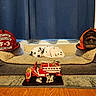 Higbee is registered to the contest to win money with this photo: dalmatian, puppy, dog_bed, firefighter_helmet, fire_dept, toy_fire_truck, dalmatian_figurine, sleeping, cozy, indoor, carpet, curtains, wood_floor, decor, resting, pet, animal, firefighter_theme, red, orange