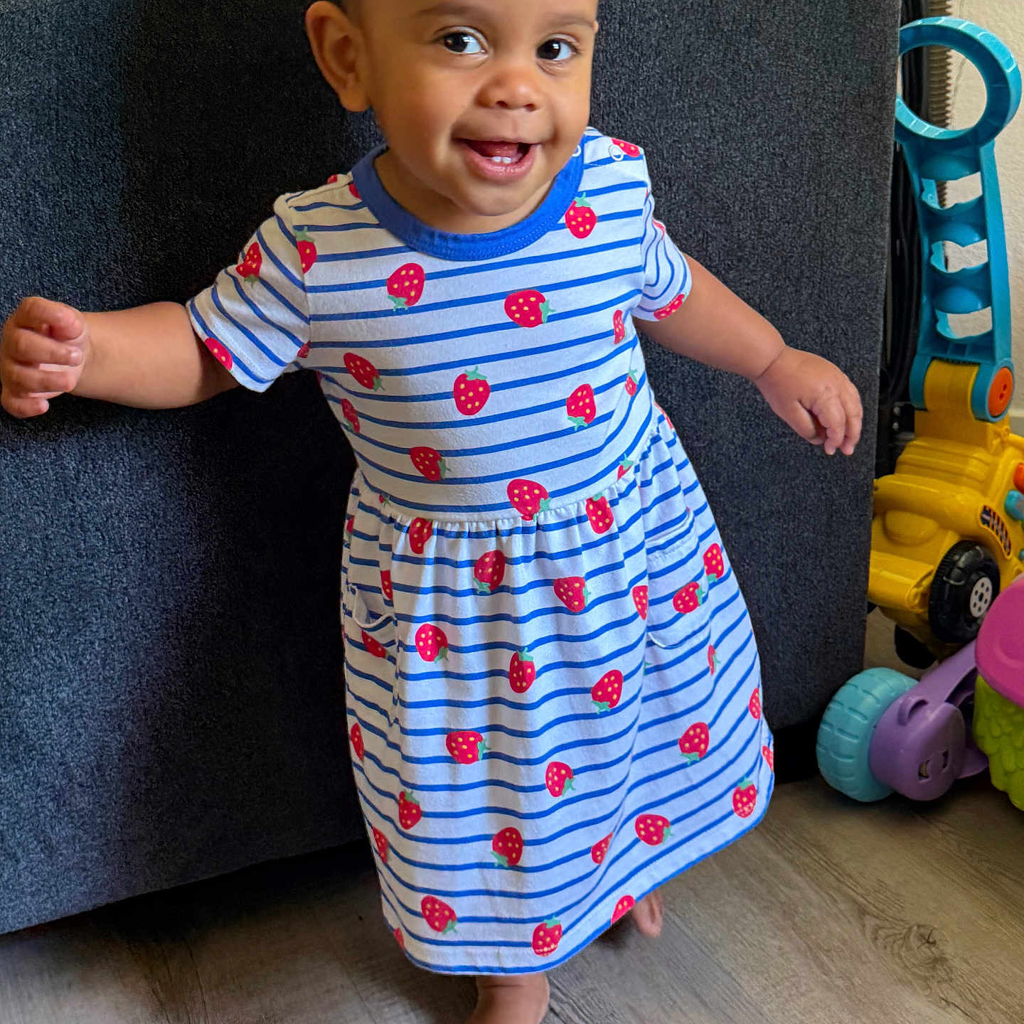 Harley Marie is registered to the contest to win money with this photo: toddler, child, smiling, dress, striped_dress, strawberry_pattern, barefoot, indoor, wood_floor, couch, toy, colorful_toys, happy, walking, person, young_child, playful, home, cute, portrait