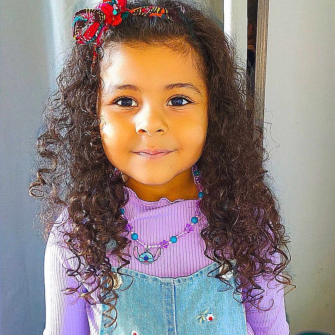 Leyah participe au concours pour gagner de l'argent avec cette photo : accessory, background, bow, child, colorful, curly_hair, cute, denim_overalls, fashion, flower_embroidery, girl, happy, indoors, necklace, person, portrait, purple_top, smiling, standing, young