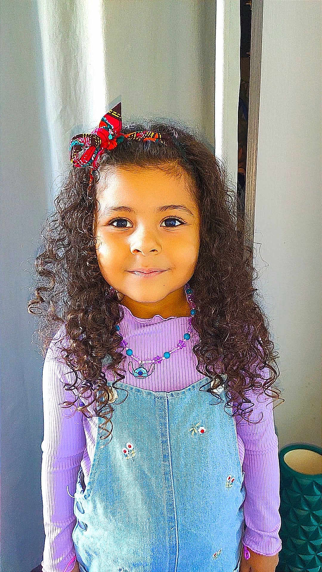 Leyah participe au concours pour gagner de l'argent avec cette photo : child, girl, curly_hair, bow, purple_top, denim_overalls, flower_embroidery, necklace, smiling, portrait, indoors, cute, young, person, fashion, accessory, happy, standing, background, colorful
