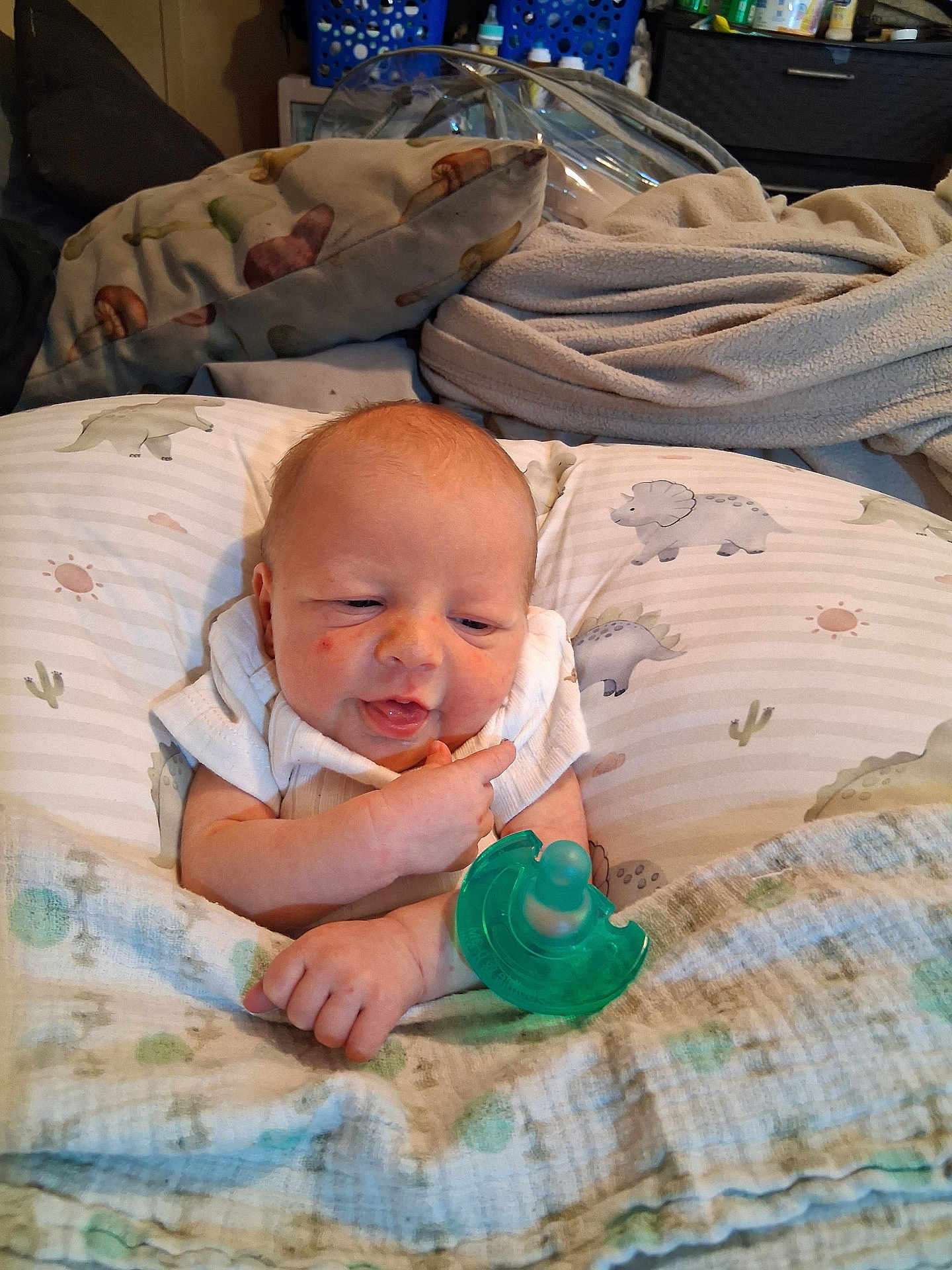 Uriah is registered to the contest to win money with this photo: baby, infant, pacifier, blanket, pillow, dinosaur_pattern, clothing, face, hand, indoor, bedroom, cute, expression, newborn, resting, cozy, portrait, child, person, soft_fabric