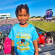 Zi'Sha is registered to the contest to win money with this photo: child, smiling, portrait, stadium, graduation, graduate, sister, blue_shirt, outdoor, sunny, crowd, spectators, scoreboard, sports_field, bleachers, sky, family_event, young_person, happy, standing