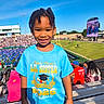 child, smiling, portrait, stadium, graduation, graduate, sister, blue_shirt, outdoor, sunny, crowd, spectators, scoreboard, sports_field, bleachers, sky, family_event, young_person, happy, standing