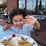 Caius joined the competition — help win amazing prizes! child, curly_hair, smiling, dessert, whipped_cream, chocolate_syrup, sprinkles, cherry, plate, table, outdoor, cafe, brick_wall, metal_chair, person, happy, food, ice_cream, playful, daylight