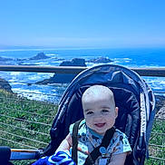 Jamison is registered to the contest to win money with this photo: baby, smiling, stroller, outdoor, ocean, cliff, blue_sky, child, happy, infant, nature, grass, sunlight, seatbelt, portrait, daylight, vacation, scenic, water, rock