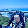 baby, smiling, stroller, outdoor, ocean, cliff, blue_sky, child, happy, infant, nature, grass, sunlight, seatbelt, portrait, daylight, vacation, scenic, water, rock