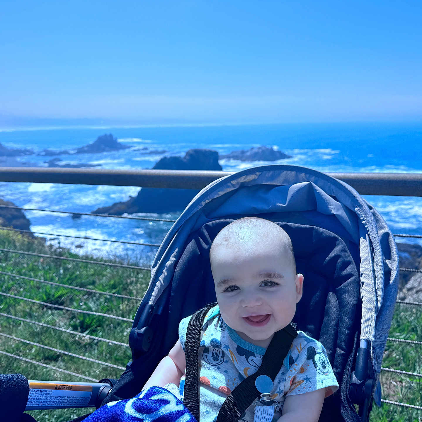 Jamison is registered to the contest to win money with this photo: baby, smiling, stroller, outdoor, ocean, cliff, blue_sky, child, happy, infant, nature, grass, sunlight, seatbelt, portrait, daylight, vacation, scenic, water, rock