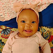 Serenity is registered to the contest to win money with this photo: baby, infant, smiling, headband, bow, pink_clothing, blanket, floral_pattern, textured_blanket, cute, happy, child, portrait, lying_down, indoors, closeup, face, person, apparel, young_child