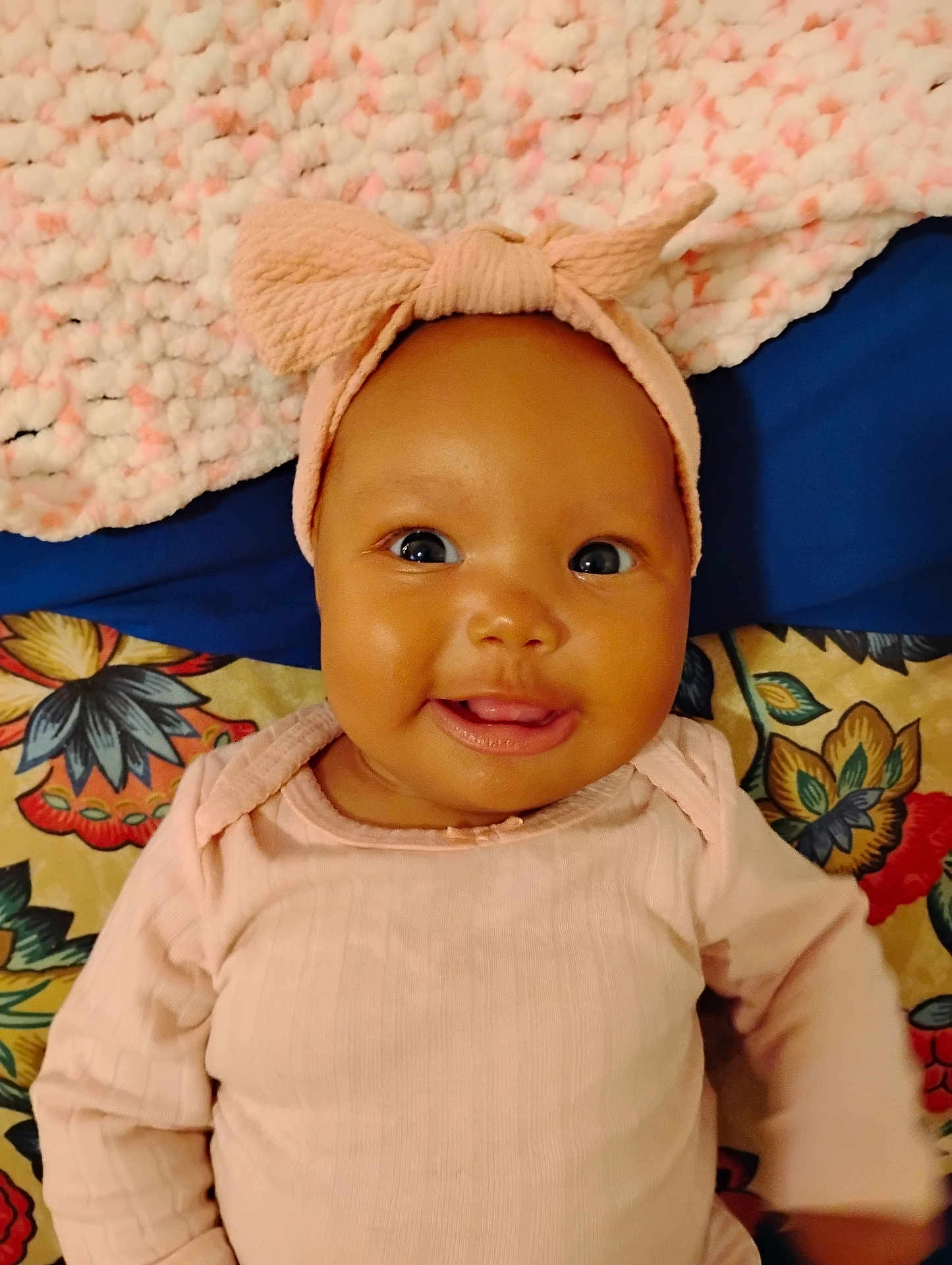 Serenity is registered to the contest to win money with this photo: baby, infant, smiling, headband, bow, pink_clothing, blanket, floral_pattern, textured_blanket, cute, happy, child, portrait, lying_down, indoors, closeup, face, person, apparel, young_child
