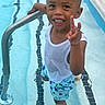 child, boy, swimming_pool, water, tank_top, swim_trunks, smiling, peace_sign, summer, outdoor, metal_railing, barefoot, happy, young, playful, casual_clothing, recreation, fun, light, daylight