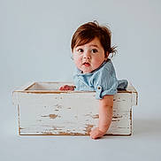 Gael is registered to the contest to win money with this photo: baby, child, blue_clothing, wooden_box, curious, innocence, portrait, toddler, studio, minimalist, soft_lighting, rosy_cheeks, short_hair, casual_wear, cute, peeking, white_background, hands, face, expression