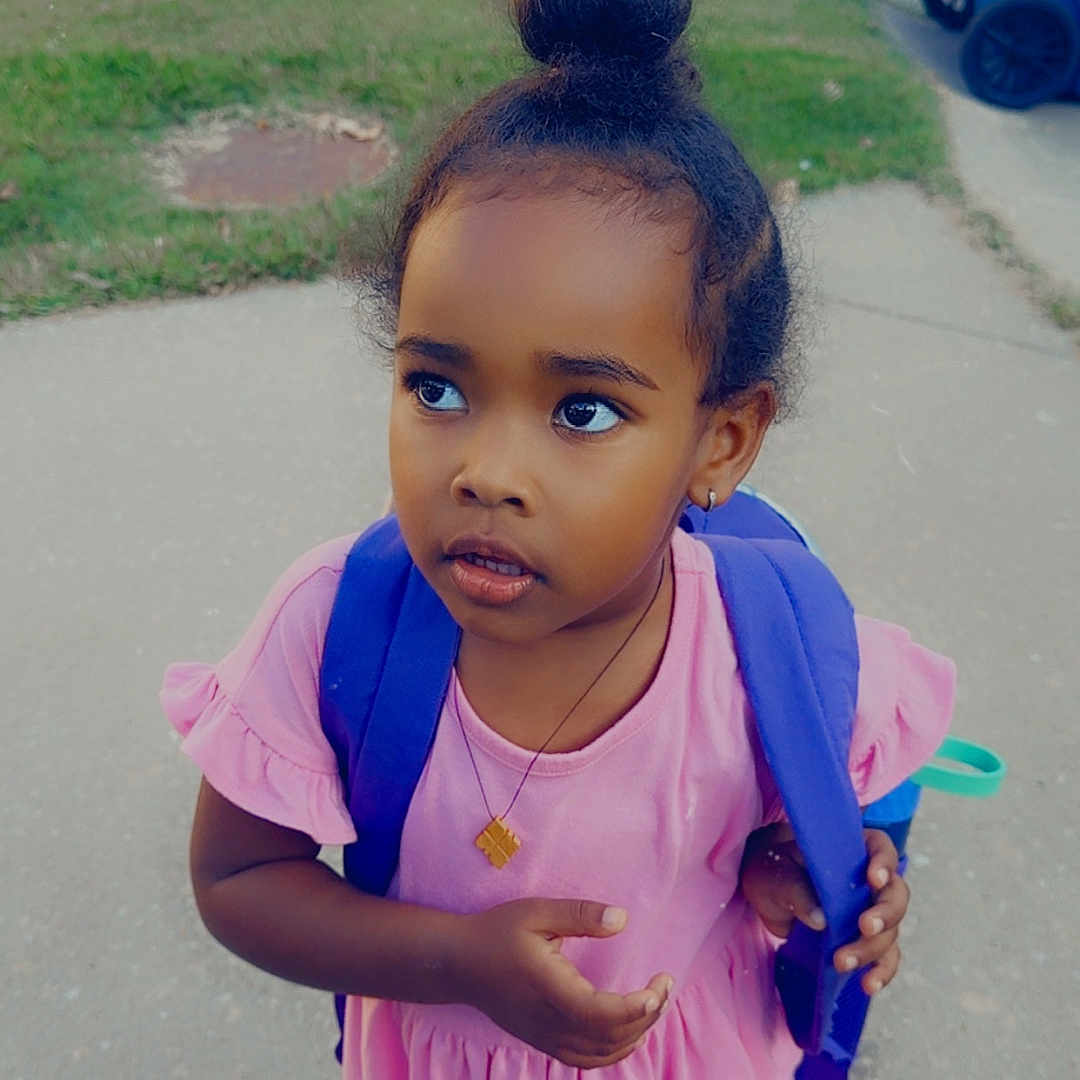 Bethlehem is registered to the contest to win money with this photo: child, toddler, girl, portrait, outdoor, sidewalk, grass, backpack, pink_dress, hair_bun, looking_away, standing, neighborhood, residential, trash_bin, blue_bin, daytime, pavement, necklace, street_photography
