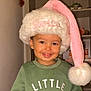 child, clothing, cozy, cute, face, festive, garland, green_sweatshirt, happy, headwear, holiday, indoor, person, plant, portrait, santa_hat, shelf, smiling, toddler, young