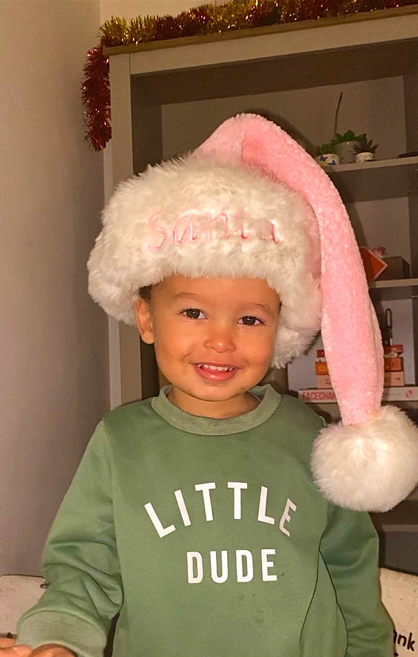 Dion is registered to the contest to win money with this photo: child, clothing, cozy, cute, face, festive, garland, green_sweatshirt, happy, headwear, holiday, indoor, person, plant, portrait, santa_hat, shelf, smiling, toddler, young