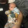 Ezra is registered to the contest to win money with this photo: baby, adult, toy, elephant_toy, blanket, bed, pacifier, clothing, hand, phone, beard, person, sleepwear, gray_blanket, comfort, indoor, rest, holding, infant, casual