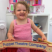 Skyla is registered to the contest to win money with this photo: blue_eyes, bookshelf, child, cute, face, furniture, hair, happy, indoor, knitted_outfit, person, pink_clothing, playroom, playtime, puppet_theatre, red_curtain, smiling, toddler, toy, wooden_sign