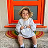 toddler, child, hat, white_clothing, purse, doorstep, window, orange_frame, smile, shoes, socks, curly_hair, colorful_rug, outdoor, person, portrait, cute, fashion, happy, sitting