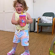 Skyla joined the competition — help win amazing prizes! toddler, child, smiling, indoor, sippy_cup, curly_hair, colorful_clothing, shoes, wooden_floor, chair, cushion, cabinet, heater, happy, person, shorts, tank_top, playful, expression, standing